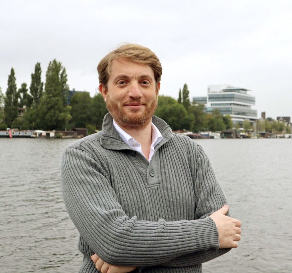 Man met grijze trui, staand aan het water met kantoorgebouwen op de achtergrond.